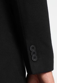 Zwarte wollen blazer manchet met drie zwarte knopen en subtiele stikdetails. Gladde textuur met een getailleerde pasvorm.