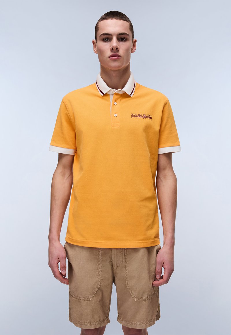 Orange polo-shirt med en hvid og maroonstribet krave, korte ærmer og broderet logo på brystet, parret med beige shorts.