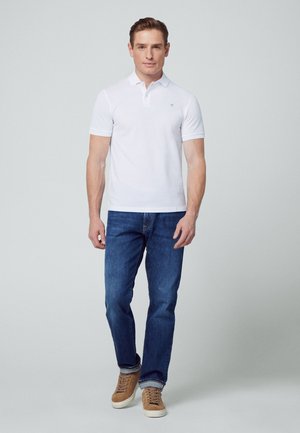 Weißes Poloshirt mit kurzen Ärmeln, blaue Jeans und beigefarbene Sneaker. Das Poloshirt hat ein kleines Emblem auf der linken Brustseite.