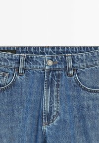 Jean en denim bleu clair avec fermeture à boutons, cinq poches et coutures visibles. Présente une légère texture délavée.