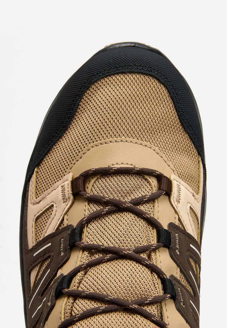 Zapato de senderismo marrón y beige con parte superior de malla texturizada, punta de goma negra y cordones marrones oscuros. Cuenta con paneles laterales reforzados.