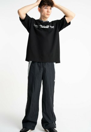 Nastolatek z krótkimi kręconymi włosami, ubrany w czarny, za duży T-shirt i luźne czarne spodnie dresowe, stojący z rękami na głowie na białym tle.