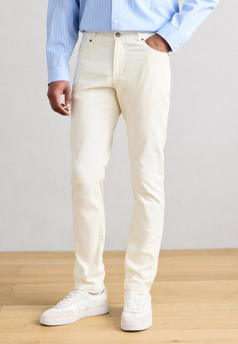 Άνδρας που φοράει μπεζ παντελόνι, λευκά αθλητικά παπούτσια και μπλε ριγέ πουκάμισο, στέκεται σε ελαφρύ ξύλινο πάτωμα μπροστά σε απλό γκρι τοίχο.