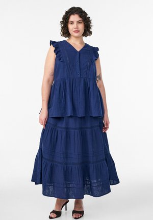 Kvinde med kort krøllet hår iført en mørkeblå flæset ærmeløs bluse og matchende lagdelt maxis skirt med sorte hælede sandaler.