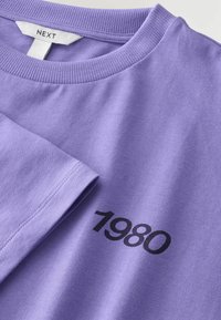 Lila Baumwoll-T-Shirt mit Rundhalsausschnitt, auf der Vorderseite mit schwarzem Aufdruck "1980". Der Stoff hat eine glatte Textur und einen gerippten Kragen.