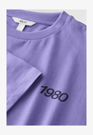 Fioletowa bawełniana koszulka z okrągłym dekoltem, na przodzie z czarnym nadrukiem "1980". Materiał ma gładką fakturę i ribbowany kołnierzyk.