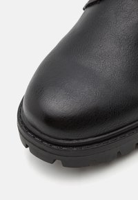 Zapato de cuero negro con punta redondeada, superficie texturizada, suela de goma robusta y costuras visibles a lo largo del borde.