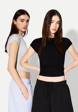 Två kvinnor bär åtsittande, kortärmade crop tops; en i grå och en i svart. Båda matchar med drawstring joggers i ljusgrå och svart.