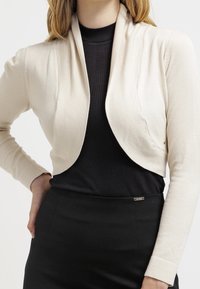 Beige zkrácený cardigan s šálovým límcem, vyrobený z měkkého pleteného materiálu. Má šikmé švy a přiléhavý design, kombinovaný s černým topem.
