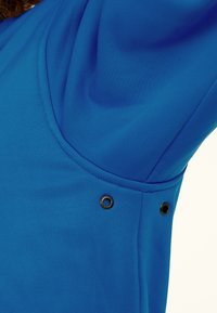 Dichtbij opname van een blauw stoffen kledingstuk met een gebogen naad en twee kleine metalen oogjes nabij de zijkant onder de arm.