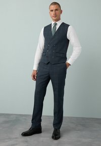 Homme portant un gilet à carreaux bleu foncé et un pantalon avec une chemise blanche, une cravate verte et des chaussures de ville noires, se tenant avec une main dans la poche.