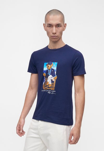 Νέος άνδρας που φοράει μπλε ναυτικό t-shirt με γραφικό ενός καθισμένου αρκούδου σε σακάκι και άσπρο παντελόνι, σε συνδυασμό με άσπρο παντελόνι σε απλό φόντο.
