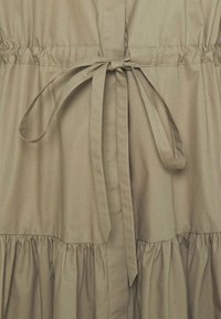 Tessuto beige con un fiocco legato al centro, con cuciture arricciate sopra e sotto il fiocco lungo la vita di un vestito o di un indumento.