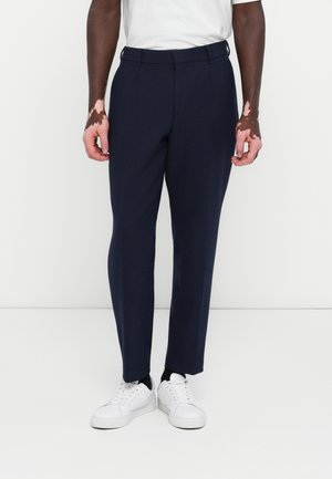 Navy tailored trousers with a smooth texture, flat front, and tapered legs. Paired with white sneakers for a contrast in color and style.