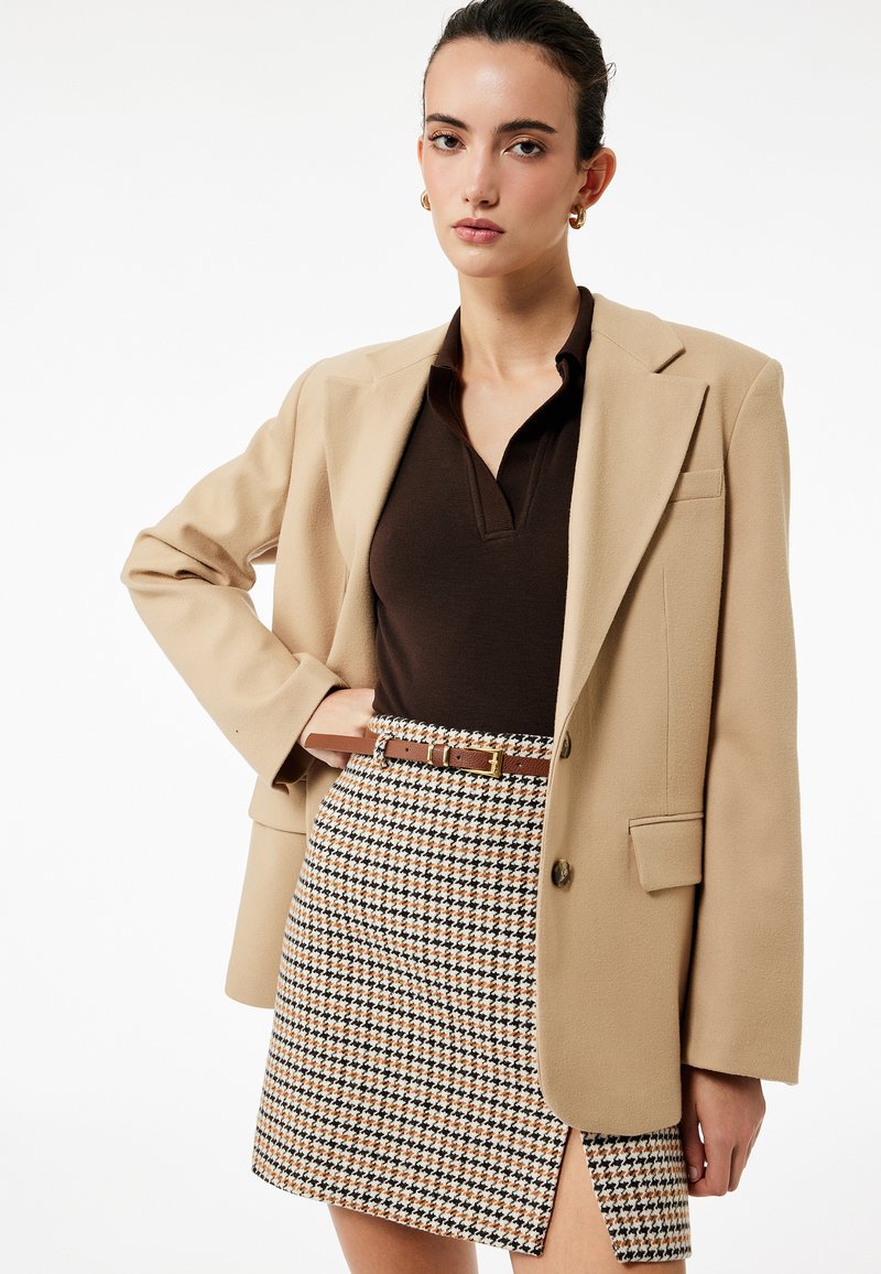 Beige blazer over en brun polo, parret med en houndstooth mini skirt i efterårsfarver og et brunt bælte. Skørtet har en sideskæring.