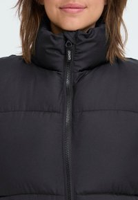 Crna puffer jakna s visokim kragnom, s prednjim zatvaračem i dizajnom s prošivenim panelima. Izrađena od glatkog, podstavljeng materijala.