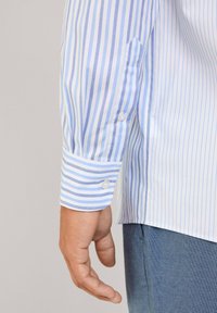 Camicia a righe blu e bianche con polsini abbottonati, caratterizzata da un design slim fit e tessuto di cotone liscio. Una mano è visibile appoggiata sulla camicia.