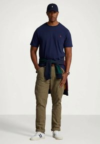 Mężczyzna w granatowej czapce, granatowej koszulce, khaki spodniach cargo, białych sneakersach i bluzie związanej w talii, stojący na jednolitym tle.