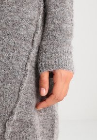 Dettaglio di un maglione grigio lavorato a maglia che presenta un tessuto morbido e testurizzato con un polsino a costine e una vestibilità rilassata, che mostra una mano sul bordo.