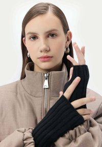 Brun zip-up jacka med hög krage, försedd med dragkedja i silverfärg. Svarta ribbade fingervantar utan fingertoppar framhäver outfitens textur.