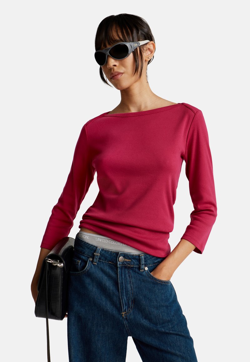 Frau mit dunklen Sonnenbrillen, rotem Langarmoberteil, blauen Jeans, die eine schwarze Handtasche trägt und mit der anderen Hand in der Tasche steckt.