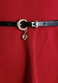 Ceinture en cuir verni noir avec une boucle ronde en argent, ornée d'un charme en forme de cœur doré, sur un fond rouge texturé.