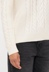 Maglione bianco a trecce con polsini e bordo a coste, caratterizzato da motivi testurizzati e una vestibilità ampia, abbinato a pantaloni in denim scuro.