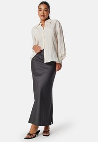 Blouse blanche texturée avec un devant à boutons ; manches longues. Jupe longue en satin gris foncé avec fente latérale et design minimaliste. Sandales à talons noires à lanières.