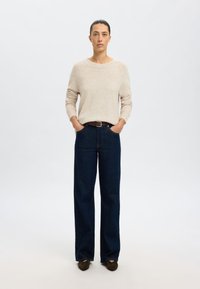 Pulover bej din lână cu mâneci lungi, asociat cu blugi largi de un albastru închis. Un brâu maro accentuează talia. Texturi netede, design simplu.