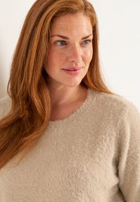 Beiger, flauschiger Pullover mit rundem Ausschnitt. Weiche Textur und leicht lockere Passform, mit einem dezenten Glanz. Haare, die über die Schultern fallen.