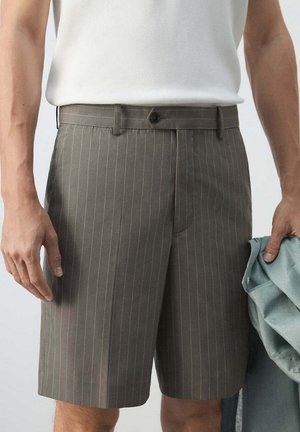 Hombre con pantalones cortos marrones con rayas finas y camisa blanca sin mangas, sosteniendo una prenda azul claro doblada en su mano derecha.