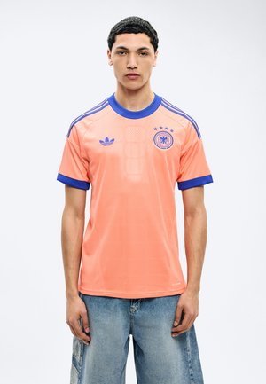 Νεαρός άνδρας που φοράει αθλητική φανέλα ροδακινί με μπλε περιθώρια και λογότυπα, στέκεται μπροστά σε απλό λευκό φόντο.