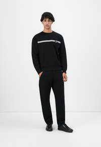 Young man standing with hands in pockets wearing black sweatshirt with white stripes, black pants, and black sneakers against white background.