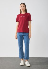 T-shirt en coton rouge à manches courtes avec le texte blanc imprimé "C'est la vie Paris", associé à un jean droit bleu et des baskets blanches.