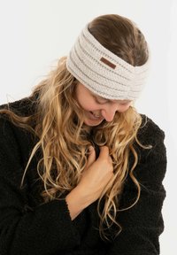 Fascia in maglia grigia chiaro, con un motivo testurizzato e un piccolo patch logo marrone. Adatta morbida ed elastica, indossata con un cappotto nero.