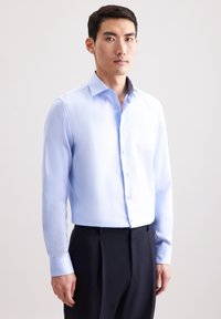 Camicia a maniche lunghe blu chiaro con chiusura a bottone, texture liscia e dettagli del colletto a fantasia in contrasto, abbinata a pantaloni scuri.