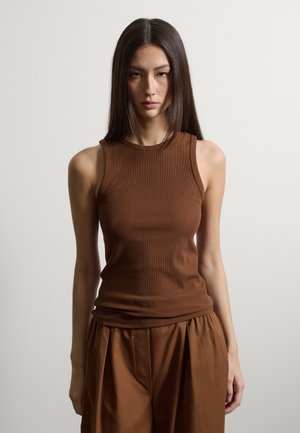 Mujer joven con cabello largo y liso de color oscuro, vestida con una blusa marrón sin mangas y pantalones marrones holgados, posando frente a un fondo claro y liso.