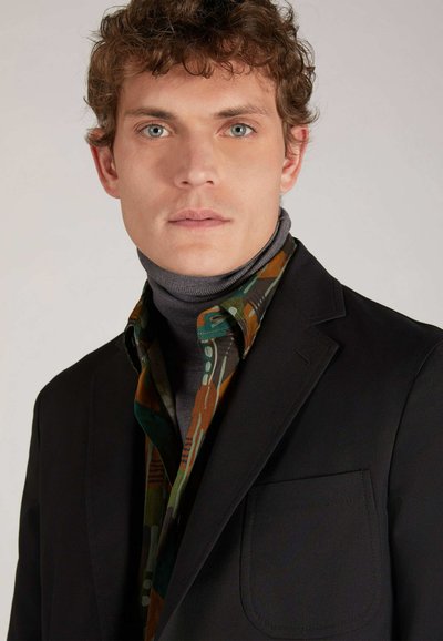 Blazer nero con una tasca, sovrapposto a una camicia a fantasia che presenta tonalità di verde, marrone e grigio, e un dolcevita grigio sottostante.