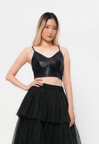 Schwarzes Leder-Crop-Top mit verstellbaren Spaghettiträgern, das über einen V-Ausschnitt und genähte Details verfügt. Kombiniert mit einem schwarzen, geschichteten Tüllrock.