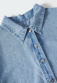 Col de chemise en denim bleu clair avec des boutons-pression métalliques visibles le long de la patte de boutonnage avant sur une surface plane.