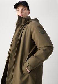 Olivgrüne wasserdichte Jacke mit Reißverschluss, großem Kapuzen und Markenzeichen auf dem linken Ärmels. Verfügt über Seitentaschen und verstellbare Bündchen.