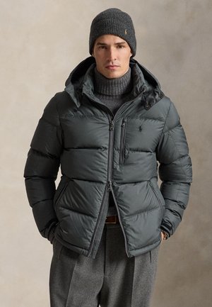 Homme portant une doudoune gris foncé, un bonnet tricoté assorti, un pull à col roulé gris et un pantalon gris, debout devant un fond neutre.