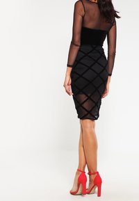 Nainen, jolla on yllä musta läpikuultava pitkähihainen paita ja ristiinkäyvä kuviollinen hame, yhdistettynä punaisiin korkeatöyhtöisiin nilkkaketjukenkiin.