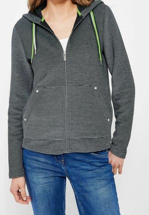Femme portant un sweat à capuche gris zippé avec des cordons verts et gris, par-dessus un haut blanc, associé à un jean bleu en denim.