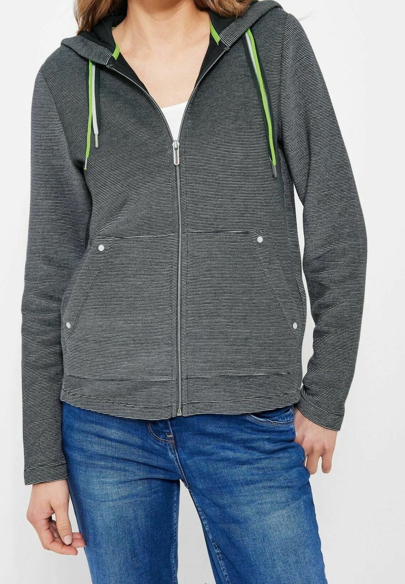 Femme portant un sweat à capuche gris zippé avec des cordons verts et gris, par-dessus un haut blanc, associé à un jean bleu en denim.