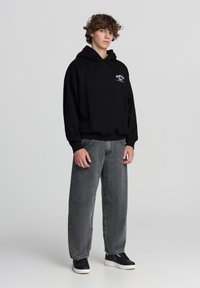 Felpa nera con cappuccio e tasca frontale, con dettagli ricamati, abbinata a jeans grigi a vestibilità ampia e sneakers nere. Design semplice e materiali.