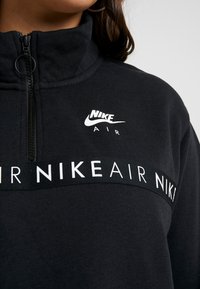 Svart Nike sweatshirt med hög krage, dragkedja och vita logotypdesigns på bröstet och bandet runt midjan.