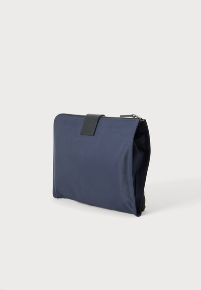 Pochette rectangulaire bleu marine avec une sangle en cuir noir et une fermeture éclair argentée, debout sur une surface gris clair.