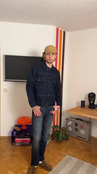 Dark denim jacket with contrasting yellow stitching, two chest pockets, paired with faded blue jeans and brown slip-on shoes.