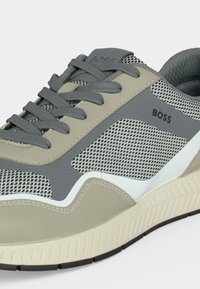 Sneakers BOSS beige et gris pour homme avec panneaux en mesh, lacets gris, col rembourré et semelle beige texturée.